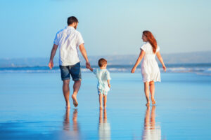 Happy family - young father, mother, baby son strolling together, child run with fun by water pool along sunset sea surf on tropical beach. Travel lifestyle, people walking with kid on summer vacation